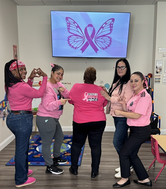 Springfield FRC staff pose in pink in front of a TV showing a butterfly and awareness ribbon. One person faces away to show the back of her shirt, which reads “Detect it, treat it, defeat it,” and others make heart shapes with their hands.